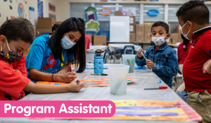 arts 4 learning miami- three students with masks on sitting at a table with a teacher, who also has a mask on, coloring large pages and sharing markers. There is a pink banner with white text on the bottom left hand side of the picture that says "program assistant".